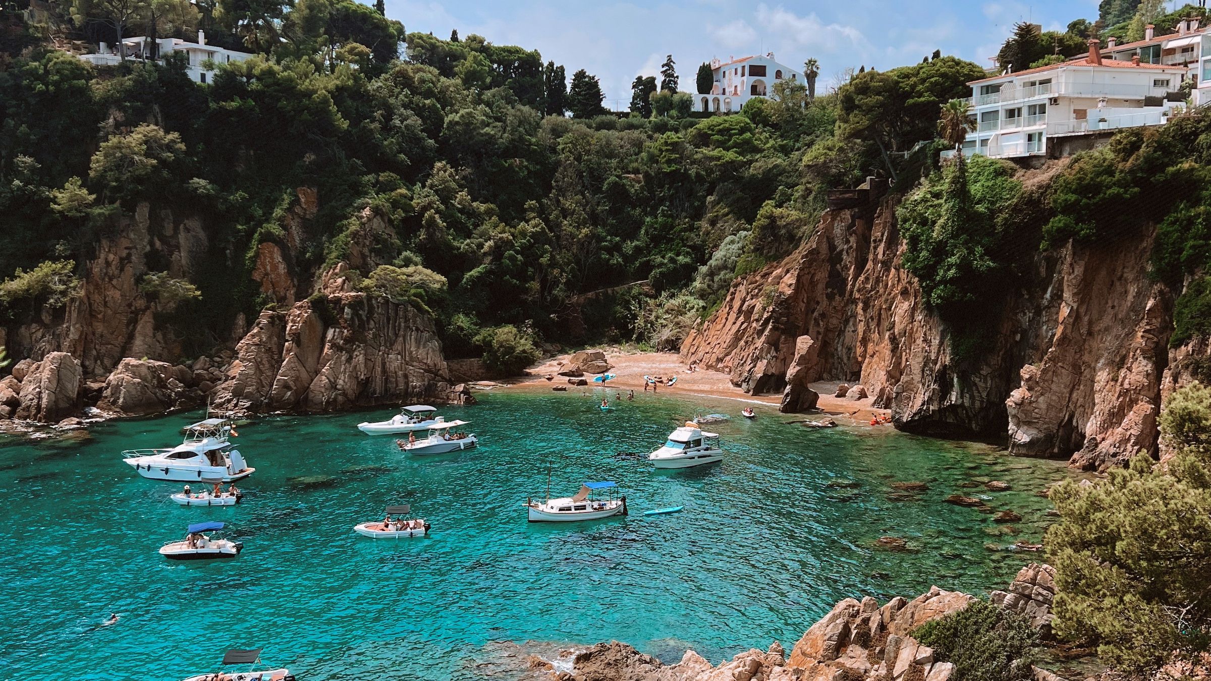 Small cove enclosed by reddish rock cliffs with clear turquoise water and pines above