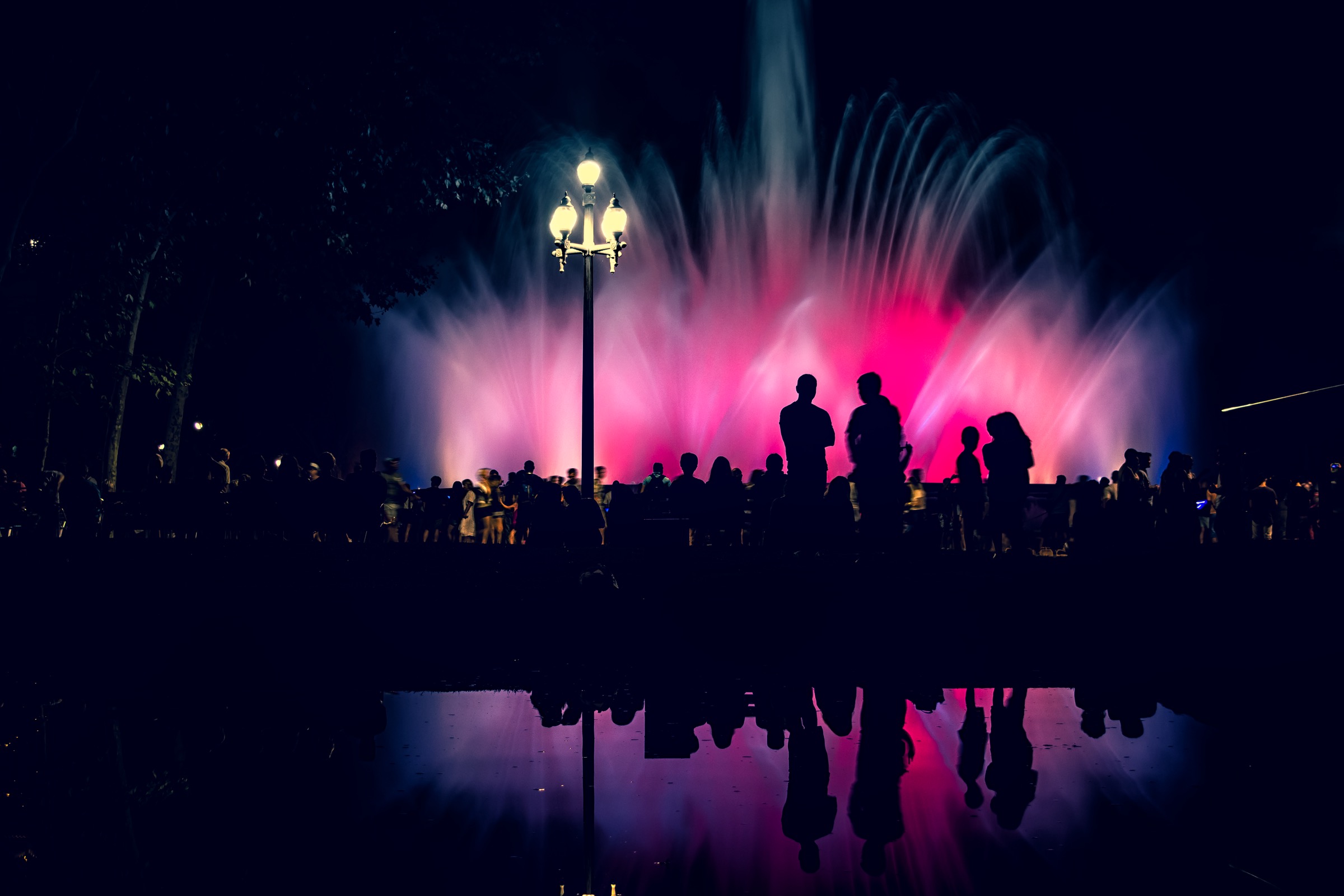 Fans of water lit magenta and blue against a night sky, an ornate cast-iron lamp post in front and a crowd watching from the railing