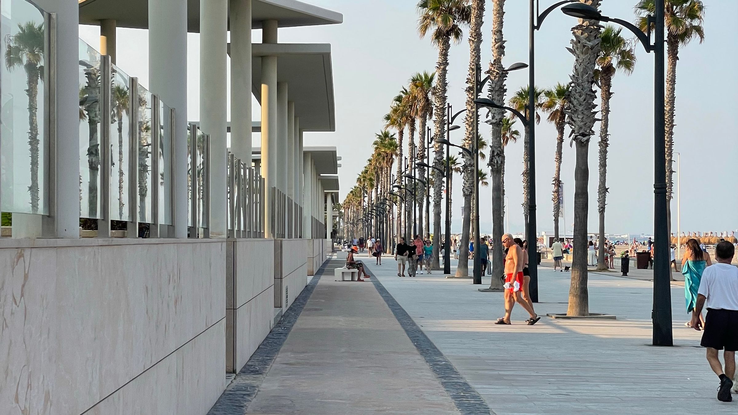 People walking along a wide palm-lined seafront promenade beside a sandy beach