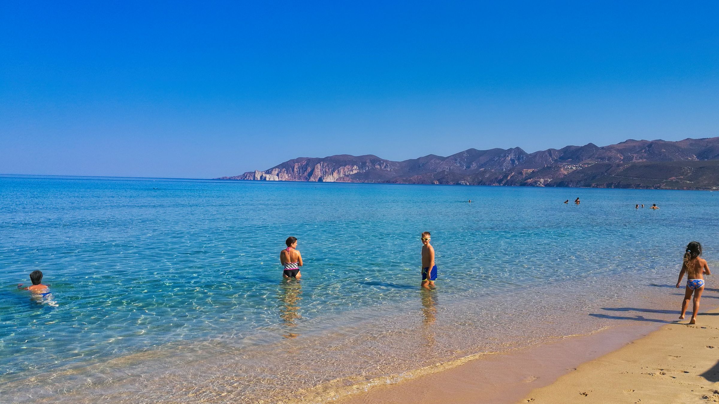 Children standing in clear shallow water at the edge of a sandy Mediterranean beach