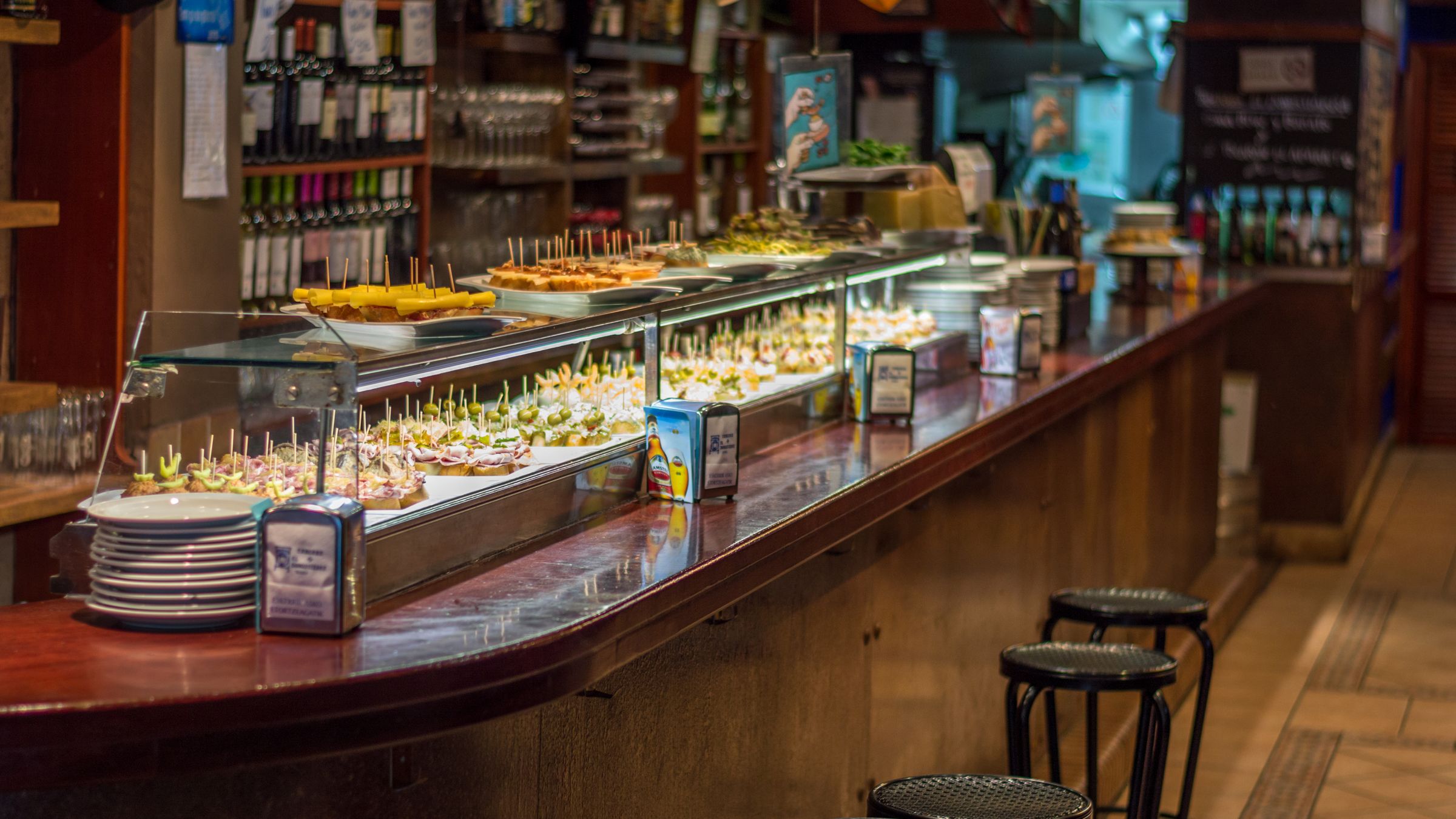 Bar counter in Spain lined with small plates of tapas behind a glass display, with wooden stools in front