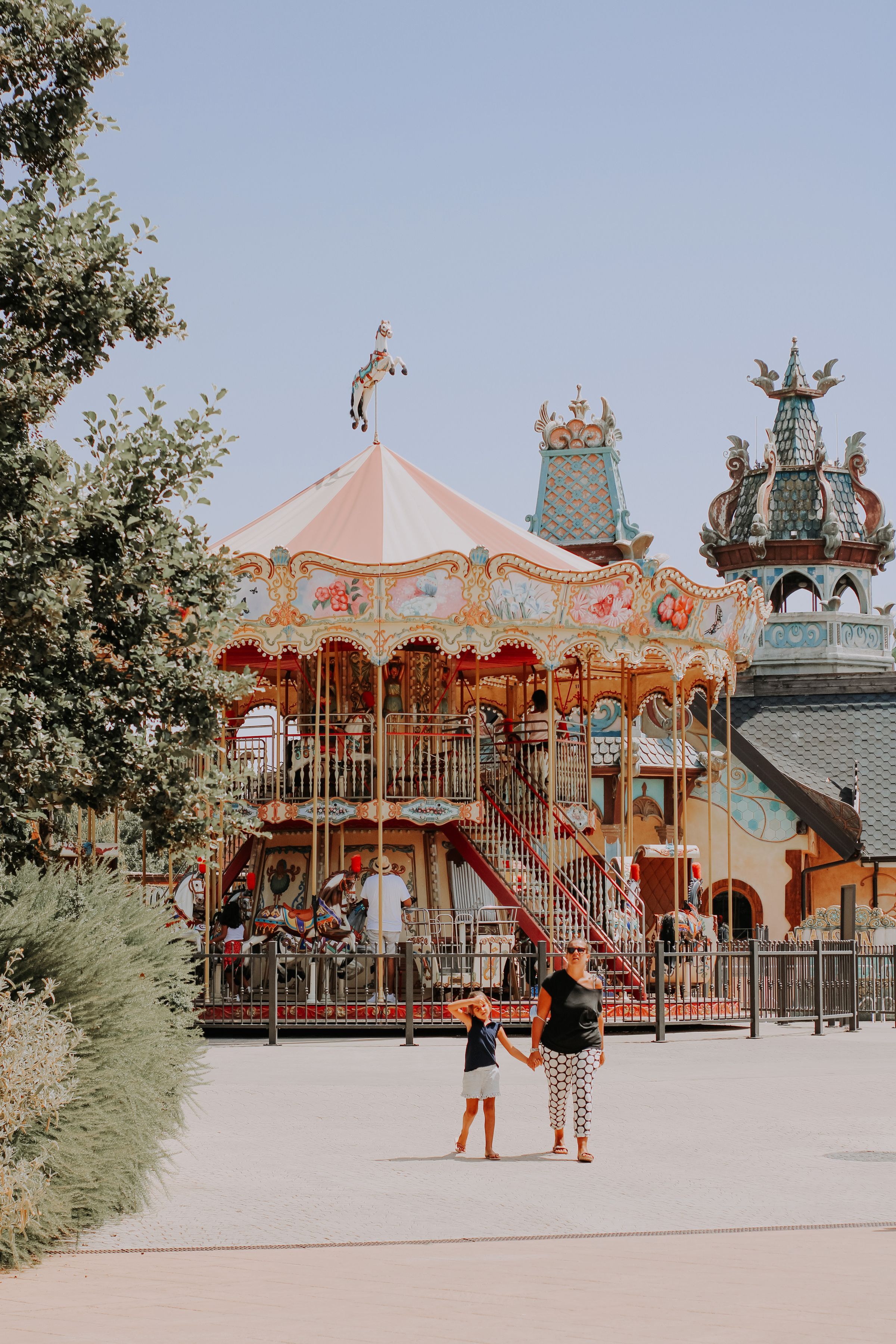 A woman and a young girl walking hand in hand across a sunlit park plaza, an ornate double-decker carousel behind them